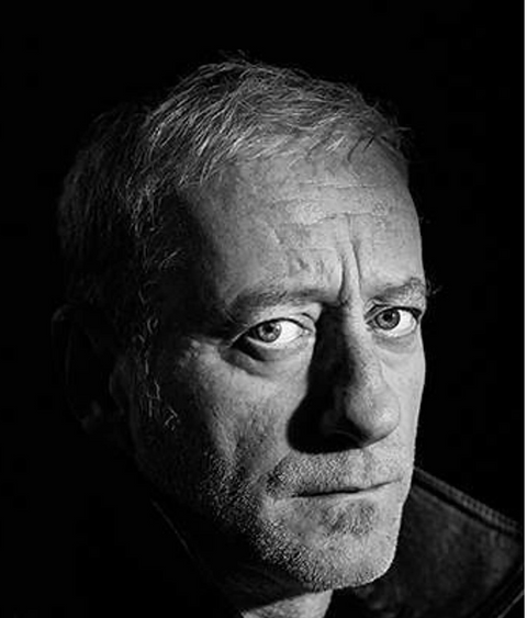 Zwart-wit portret van misdaadauteur Luc Deflo, bekend om zijn Vlaamse thrillers, met een intense blik tegen een donkere achtergrond.